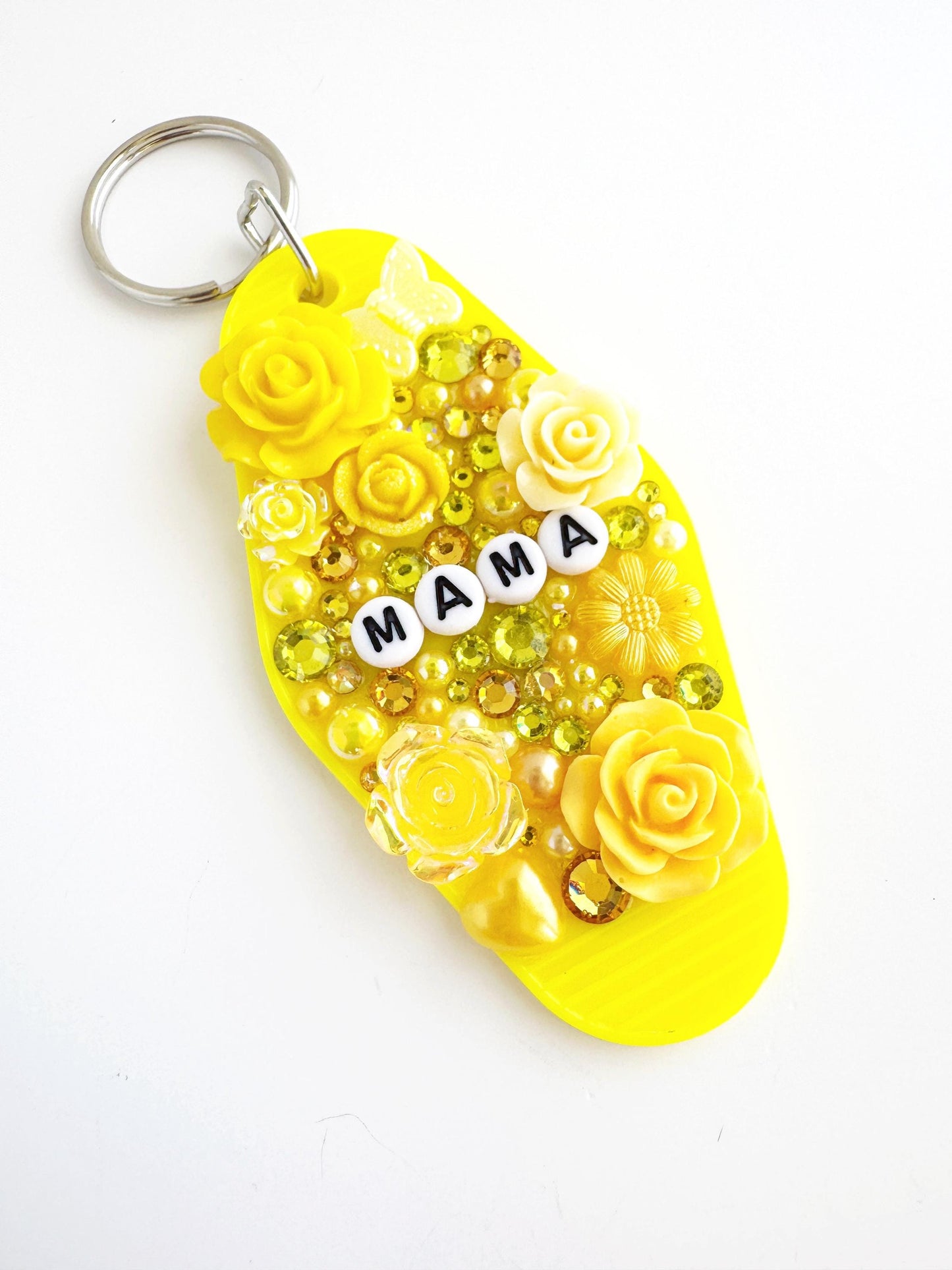 A yellow keychain with the word "MAMA" spelled out in white letters, surrounded by various decorative elements such as flowers, beads, and other embellishments.