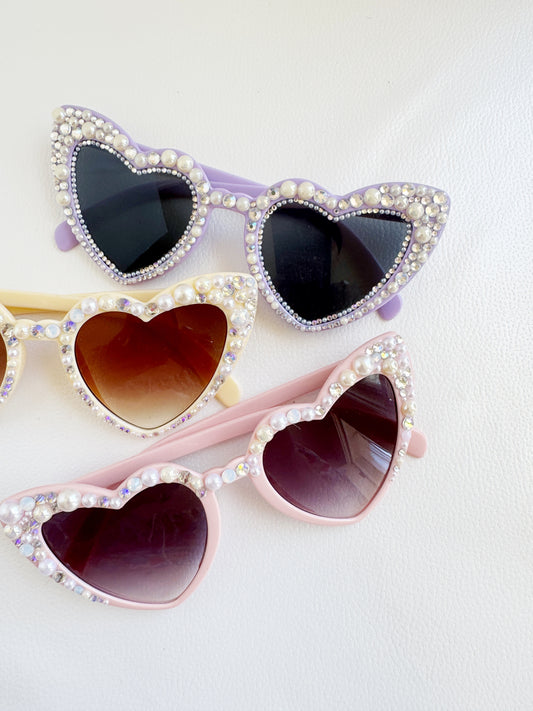 A pair of heart-shaped sunglasses with rhinestones and pearls, in various colors including purple, pink, and yellow.