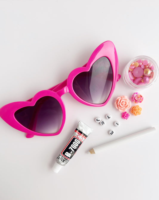 a pair of pink heart-shaped sunglasses, a tube of nail polish, and some beads, all arranged on a white background.