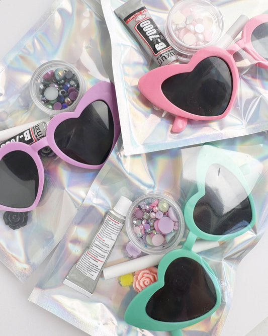 A collection of heart-shaped sunglasses, makeup items, and glittery objects arranged on a shiny, iridescent surface.