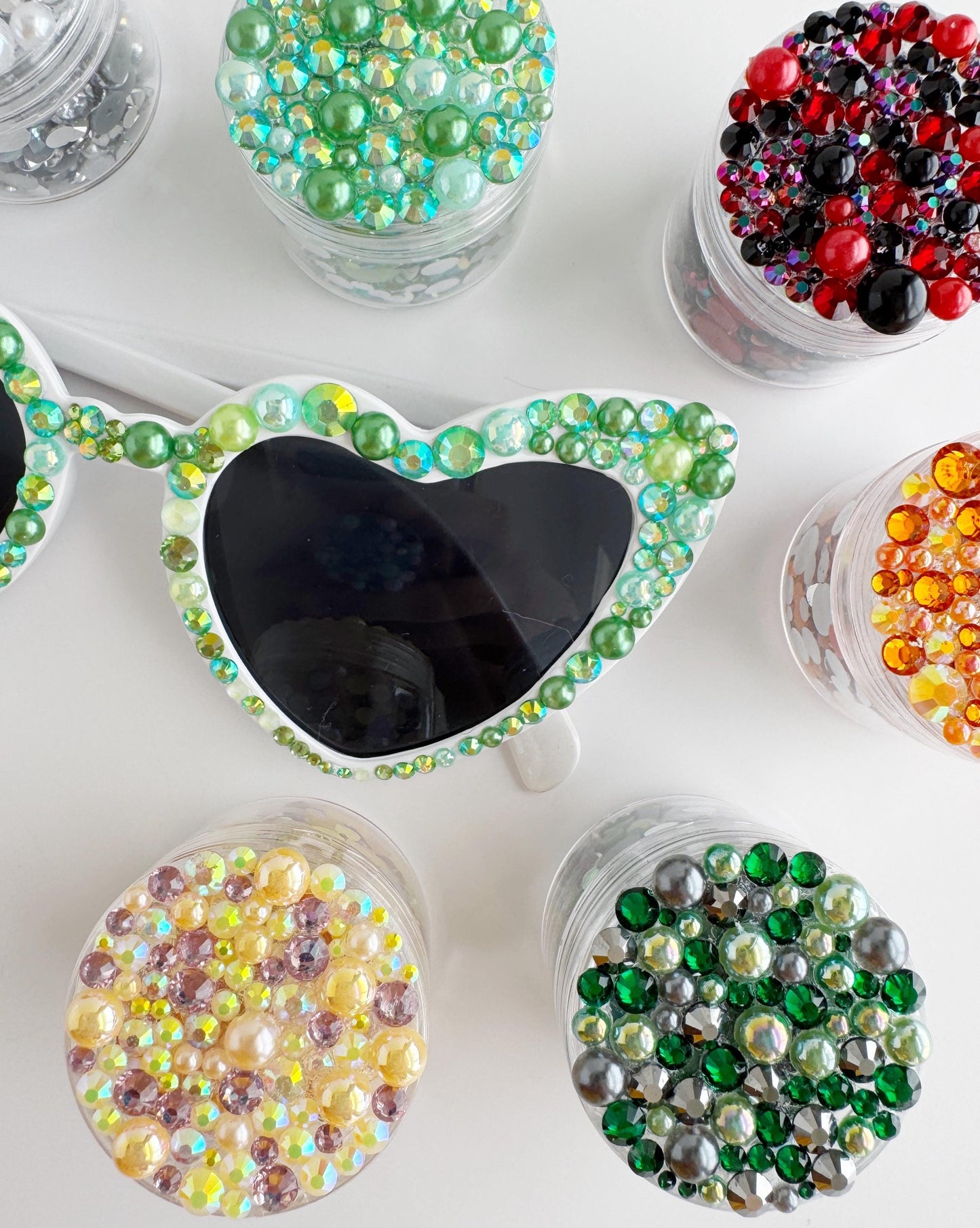 A pair of colorful, sparkling sunglasses with various colored beads and gemstones attached to them, along with other small containers filled with similar beads and gemstones.