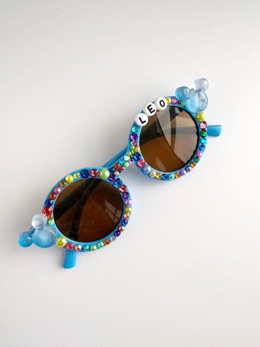 A pair of colorful, personalized sunglasses with rhinestones and the name "LEO" on the frame.