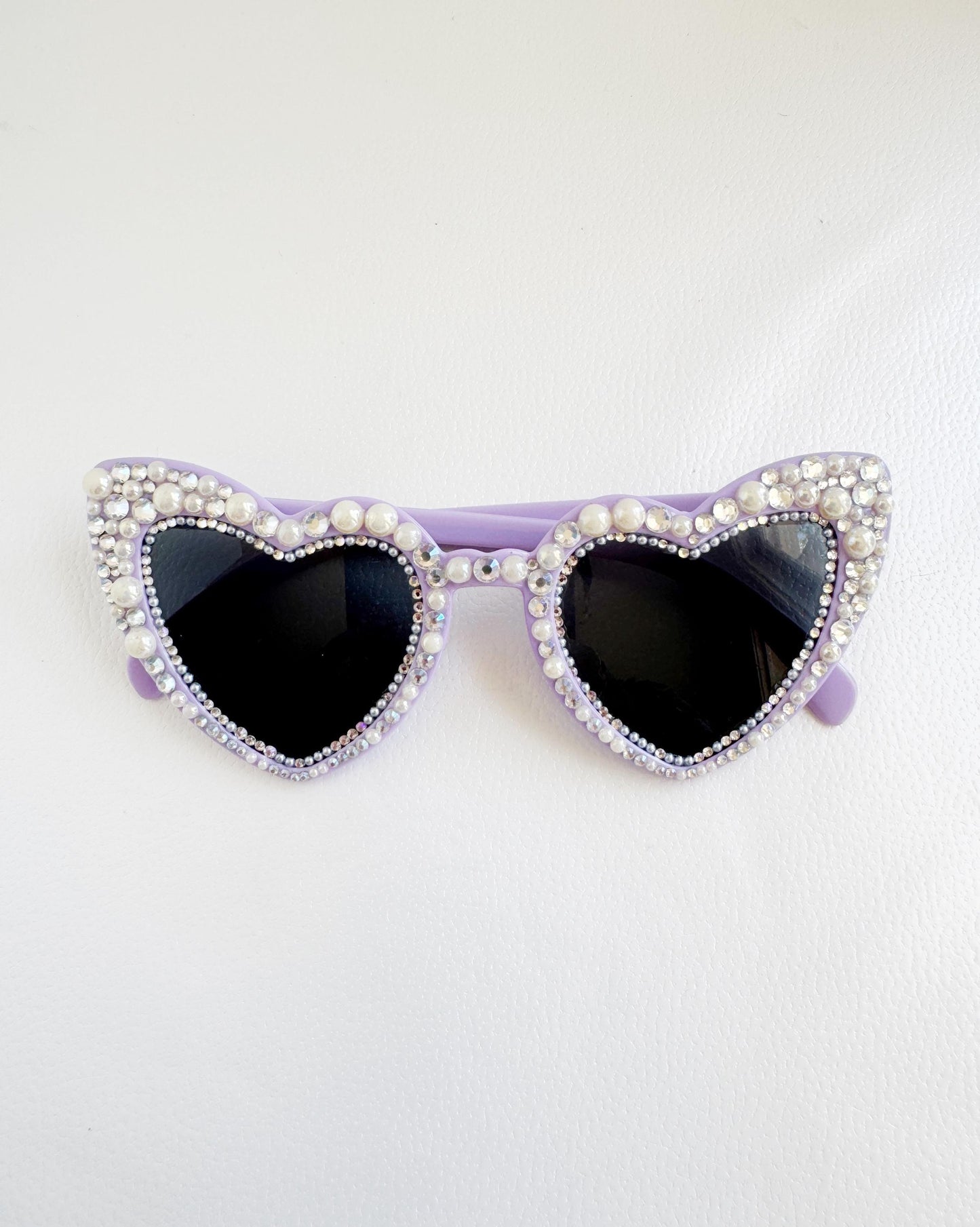 A pair of purple heart-shaped sunglasses with rhinestones and pearls on the frame.