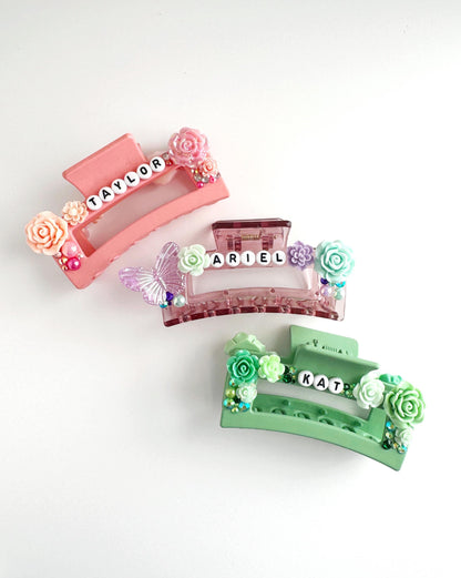 three colorful hair clips with floral decorations and the names "Taylor", "Ariel", and "Kat" written on them.