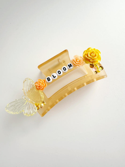 A beige hair clip with the word "BLOOM" spelled out in white letters, decorated with orange and yellow flowers and butterflies.