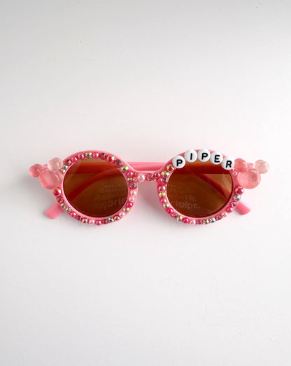 A pair of pink sunglasses with rhinestones and the word "Piper" written on them.