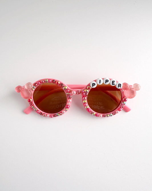 A pair of pink sunglasses with rhinestones and the word "Piper" written on them.
