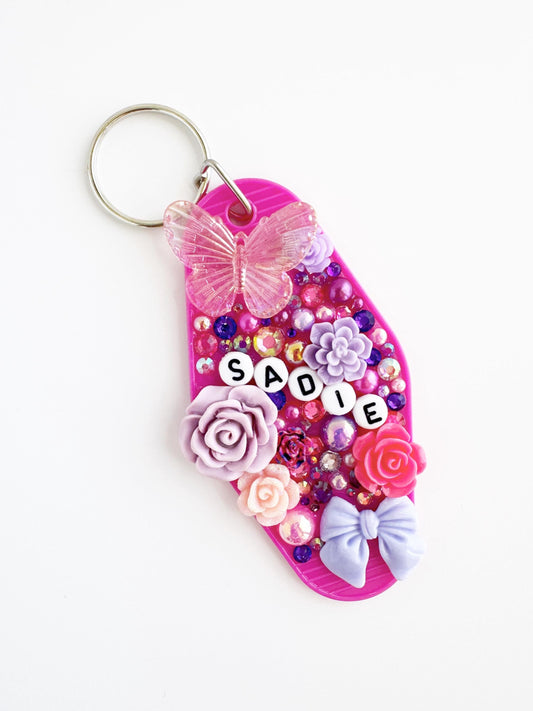 A pink keychain with various decorative elements, including a butterfly, flowers, and a name tag.