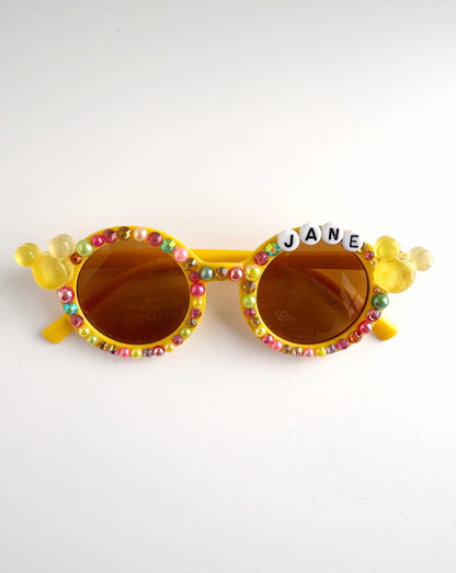 A pair of yellow sunglasses with a personalized name spelled out in beads on the frame.