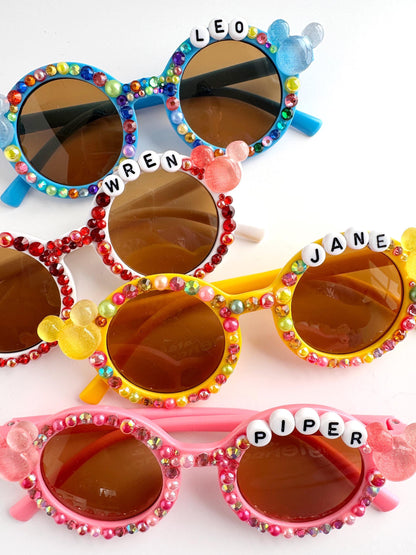 A collection of colorful, personalized sunglasses with rhinestones and name charms.