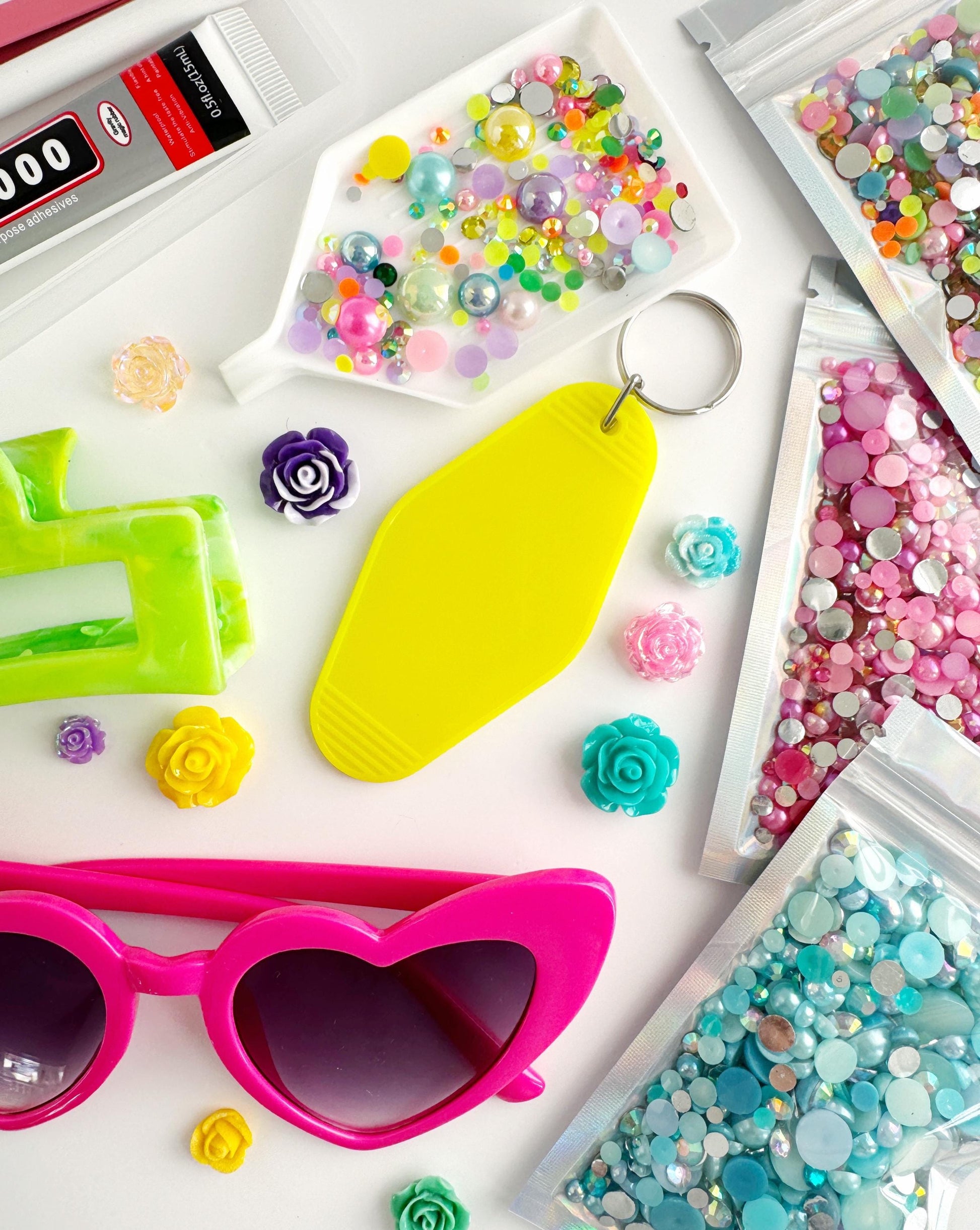A collection of colorful and creative items, including a yellow keychain, pink sunglasses, and various beads and sequins, arranged on a white background.