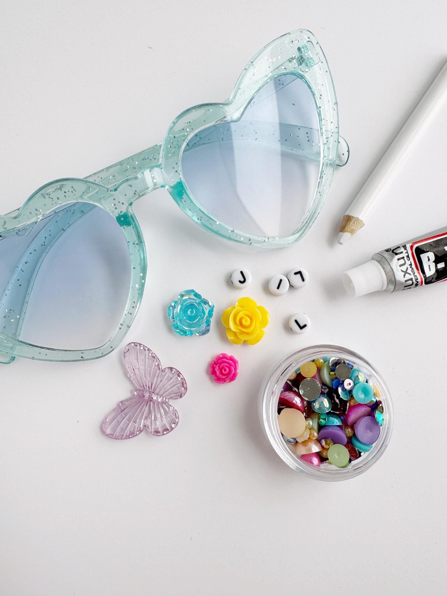 A pair of heart-shaped glasses, a small container of colorful buttons, a pen, and a small container of beads are arranged on a white surface.
