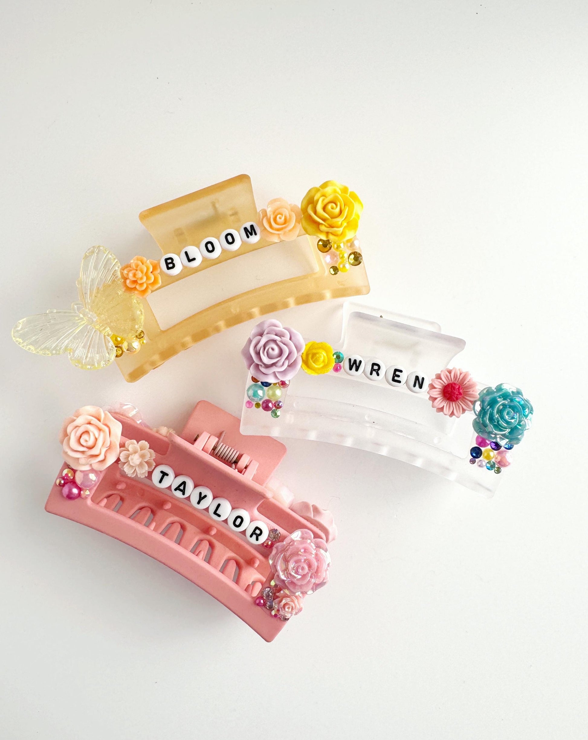 three colorful hair clips with floral decorations and the names "BLOOM", "WEN", and "TAYLOR" written on them.